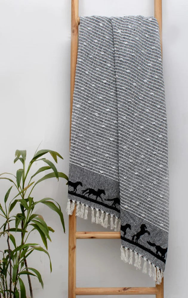 Black Cotton Woven Throw with Black Galloping Horses on Border Hangs on Wooden Ladder Against Wall. Green Plant in Corner.