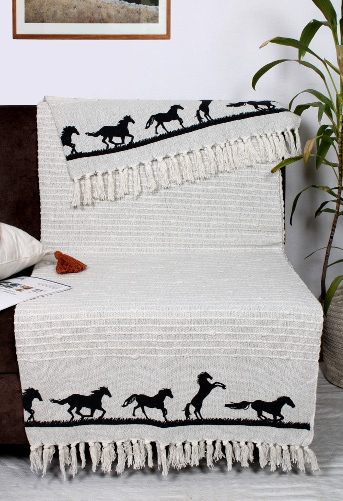 Grey Cotton Woven Throw with Black Galloping Horses on Border Spread Across Black Sofa.. Pillow and Magazine on the Sofa. Green Plant on Right Side.