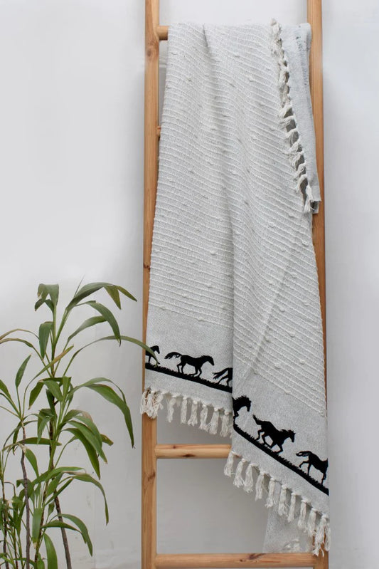 Grey Cotton Woven Throw with Black Galloping Horses on Border Hangs on Wooden Ladder Against Wall. Green Plant in Corner.