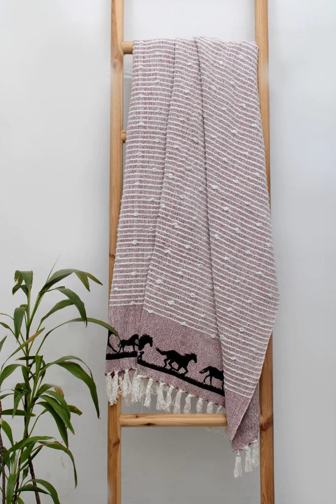Red Cotton Woven Throw with Black Galloping Horses on Border Hangs on Wooden Ladder Against Wall. Green Plant in Corner.