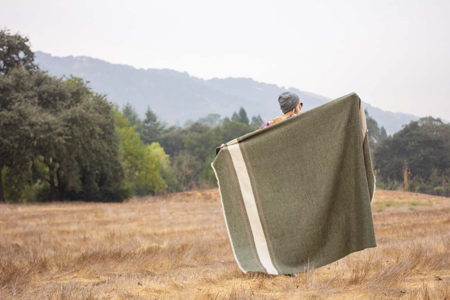 Whup Trot Travel Blanket Olive Green with Light Brown and Cream Stripes on Ends Woman Standing in Field with Blanket Spread Over Shoulders